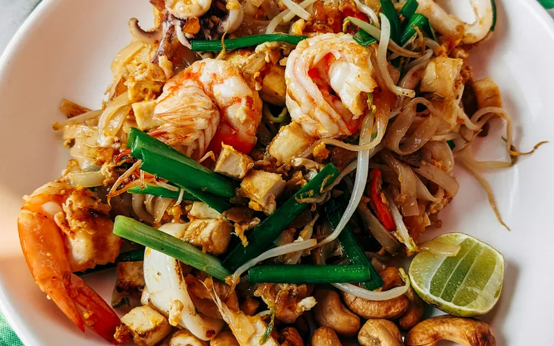 Pad Thai-style rice noodles with shrimp, calamari, golden tofu, egg, bean sprouts, green chives, roasted cashews, and a lime wedge on a white plate